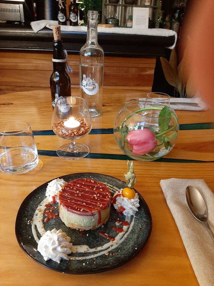 Un gâteau circulaire nappé de sauce caramel et de crème fouettée, présenté sur une assiette, elle-même posée sur une table en bois avec des boissons et une fleur.