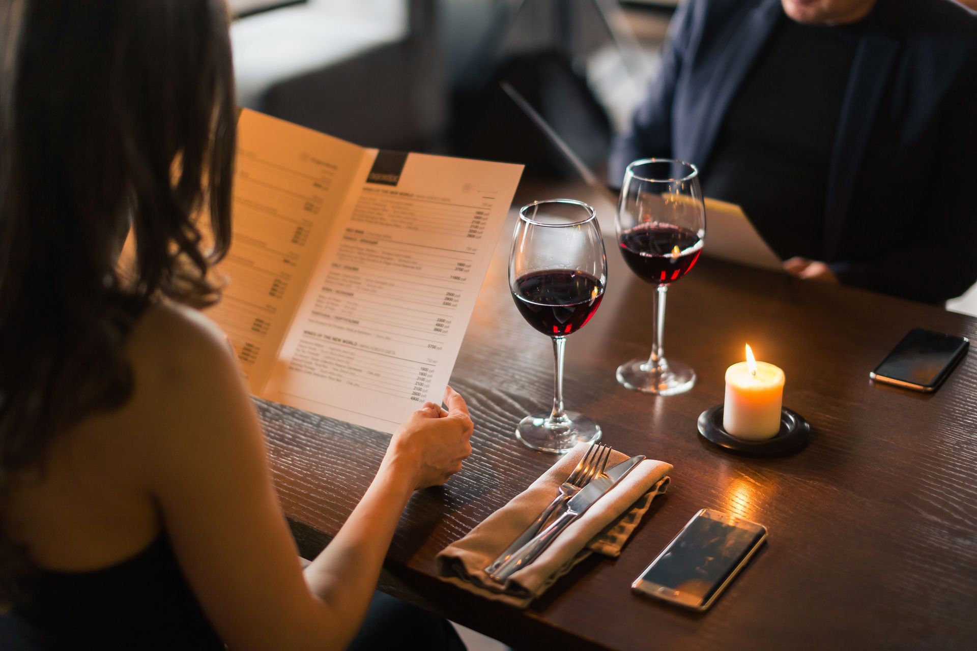 Une personne lisant un menu à une table éclairée à la bougie, avec deux verres de vin rouge et un smartphone.