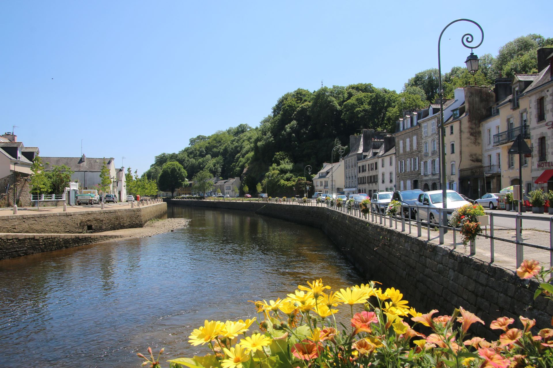 Un canal paisible traverse une petite ville française, avec ses bâtiments en pierre, ses arbres luxuriants et ses fleurs colorées au premier plan.