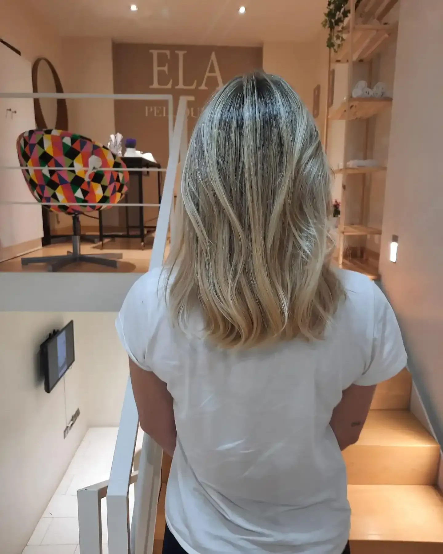 Mujer con cabello rubio en capas en un salón, de pie cerca de una escalera.