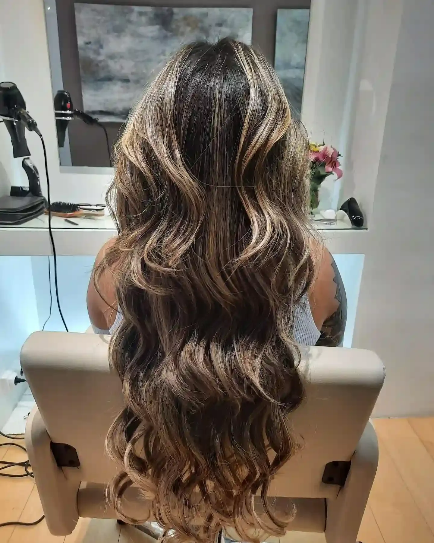 Mujer con cabello largo ondulado, con reflejos castaños y rubios, sentada en una silla de salón.