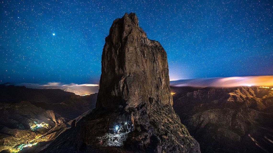 Una gran formación rocosa está rodeada de montañas por la noche bajo un cielo estrellado.