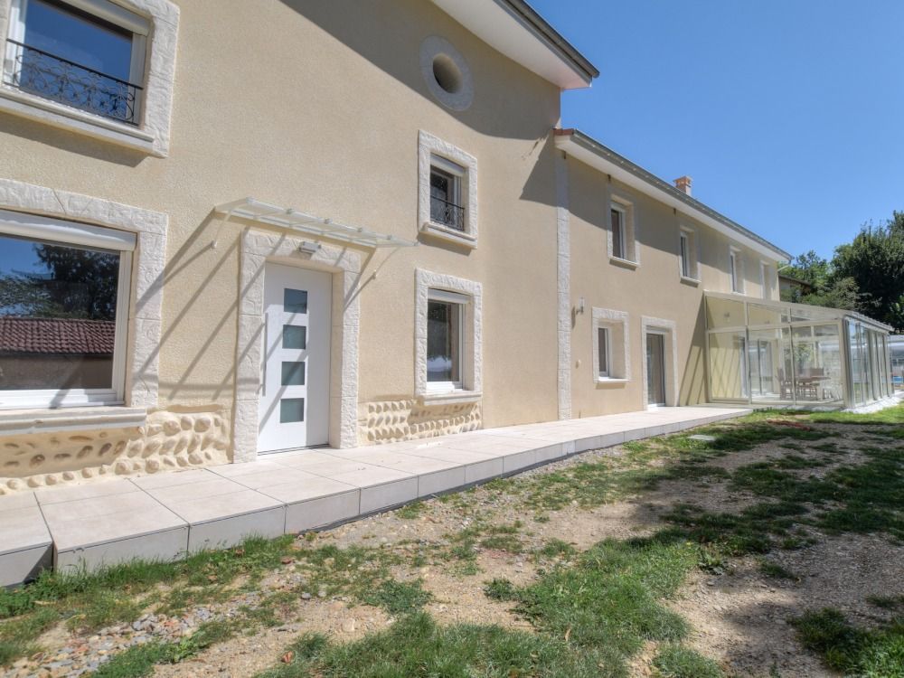 Une maison beige à deux étages avec une porte d'entrée blanche, des soubassements en pierre et un petit porche, vue depuis un jardin gazonné.