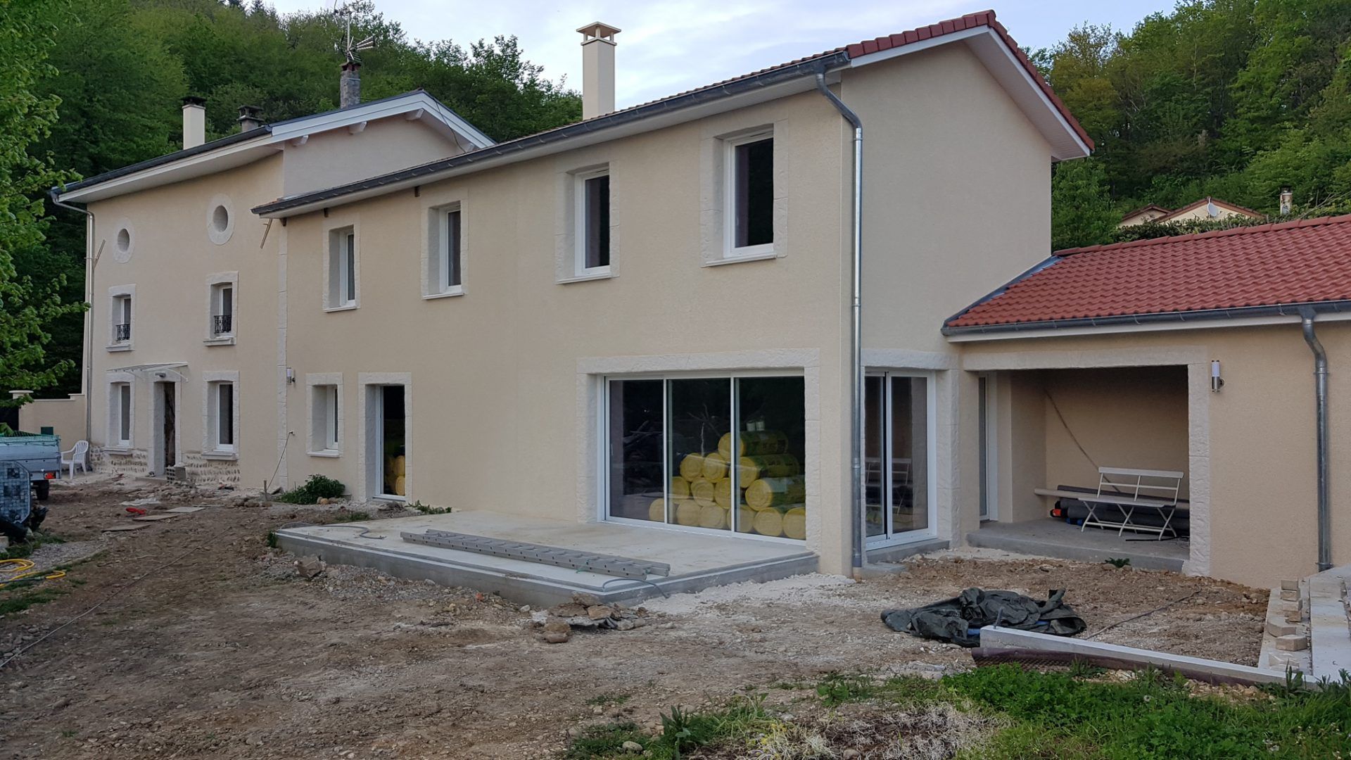 Une maison de deux étages, de couleur crème, avec une grande porte coulissante en verre, une terrasse et un garage ouvert attenant.