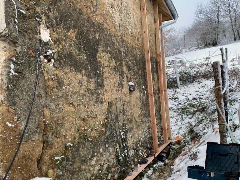 Un mur extérieur en pierre et en terre battue, patiné par le temps, en construction, avec des poutres de soutien en bois, dans un paysage rural enneigé.