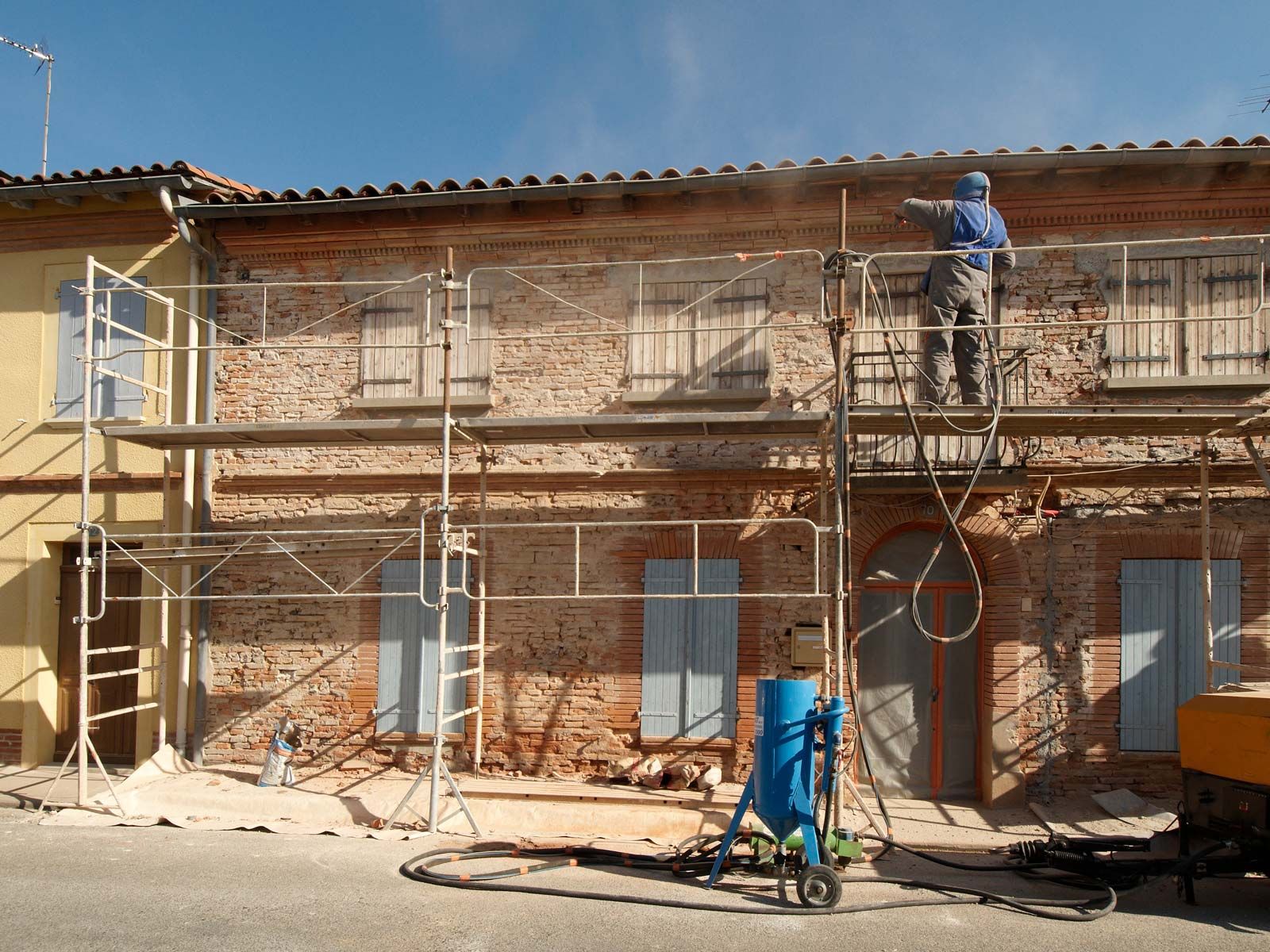 Un ouvrier sur un échafaudage utilise un outil de sablage pour nettoyer la façade en briques d'un vieux bâtiment.