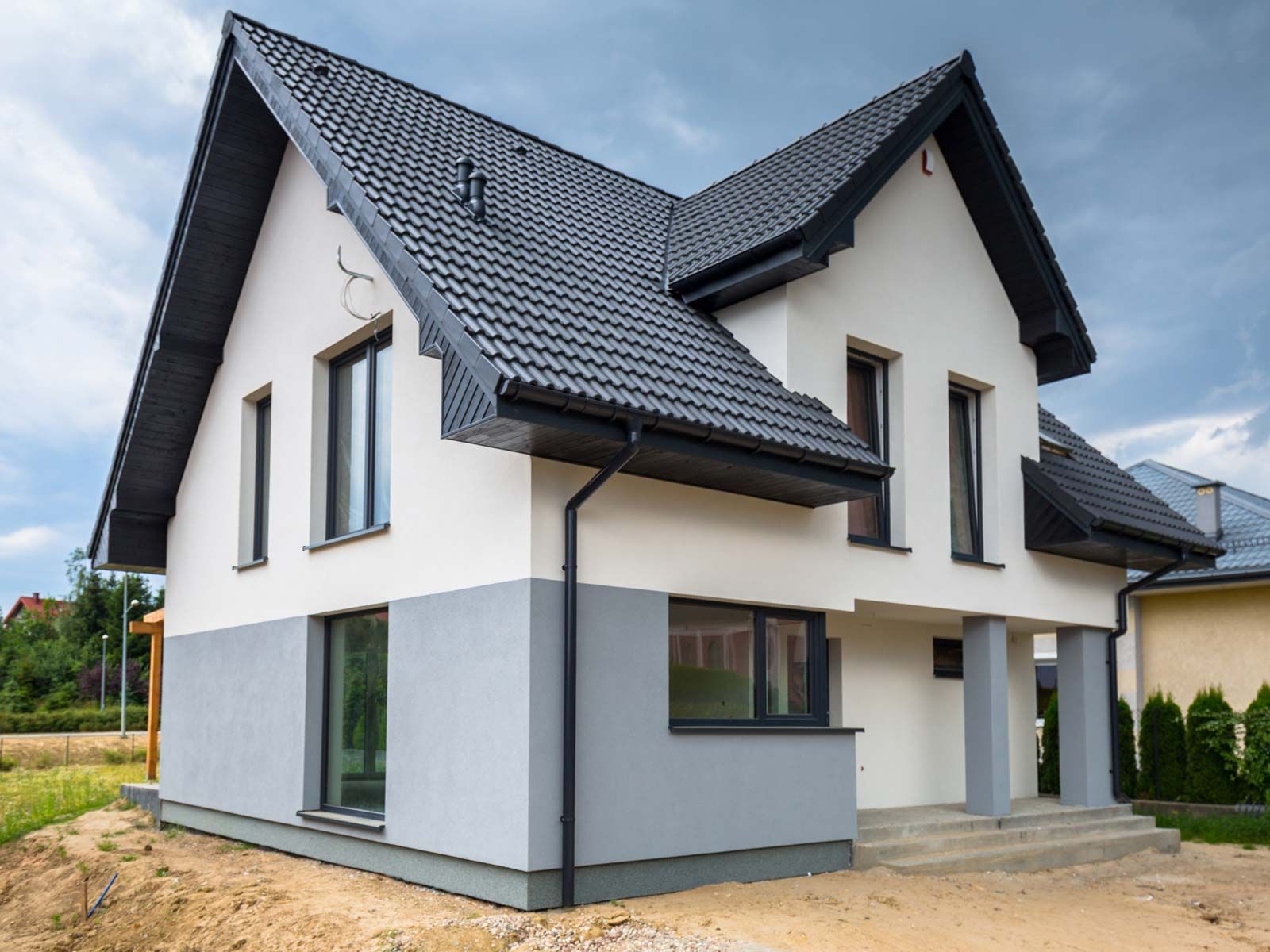 Une maison moderne de deux étages, blanche et grise, avec un toit à pignon sombre, de hautes fenêtres rectangulaires et un porche.