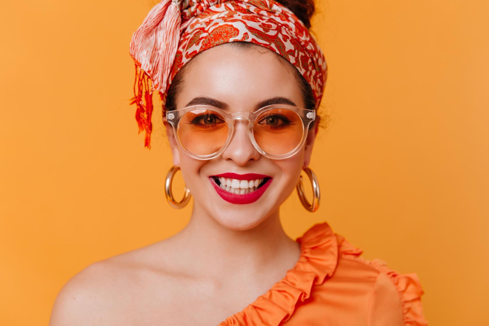 Une personne souriante portant un foulard orange, des lunettes de soleil à verres orangés, des créoles dorées et un haut orange.