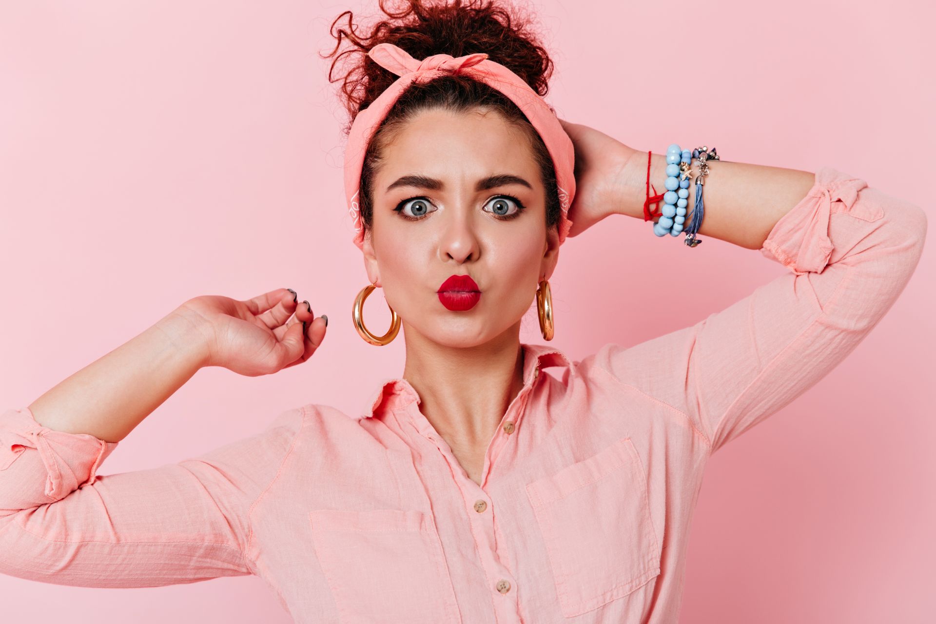 Une personne aux cheveux bouclés et portant un bandeau rose fait la moue, posant devant un fond rose et vêtue d'une chemise rose.