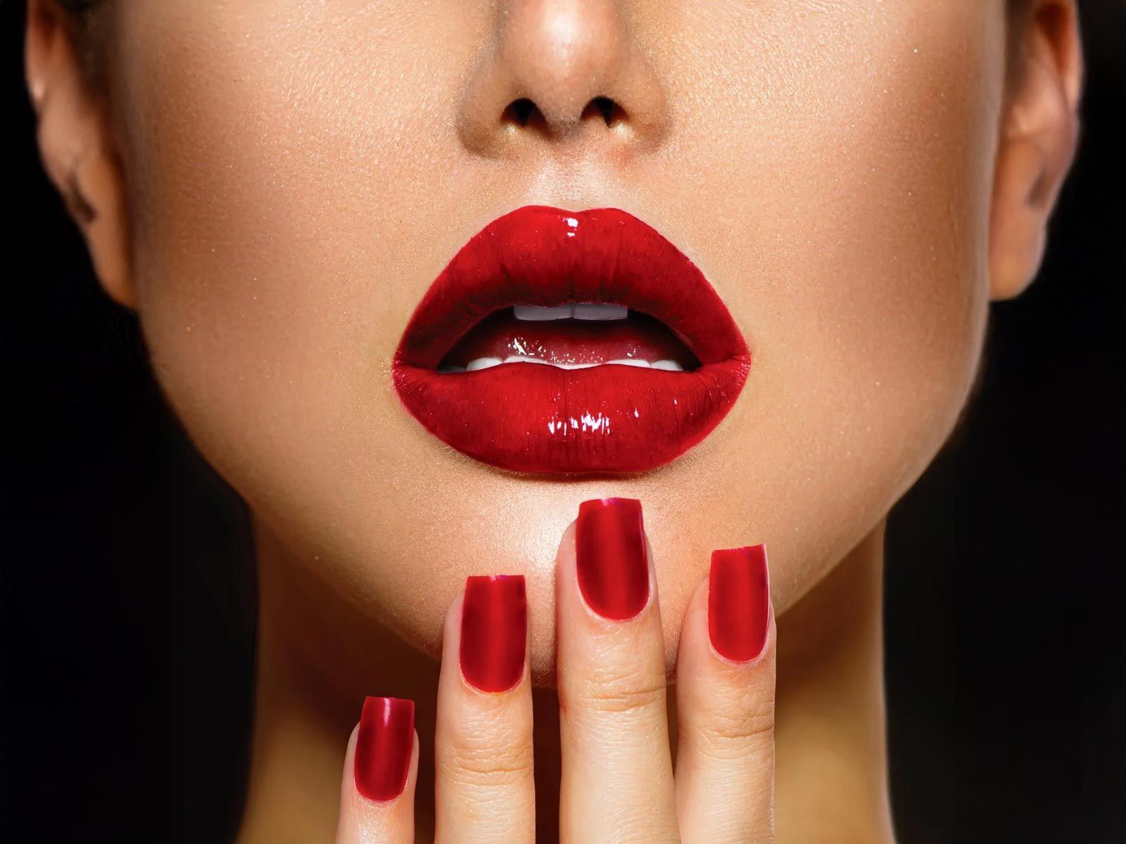 Gros plan sur des lèvres rouges brillantes et des ongles rouges assortis, posées sur le menton, sur fond noir.