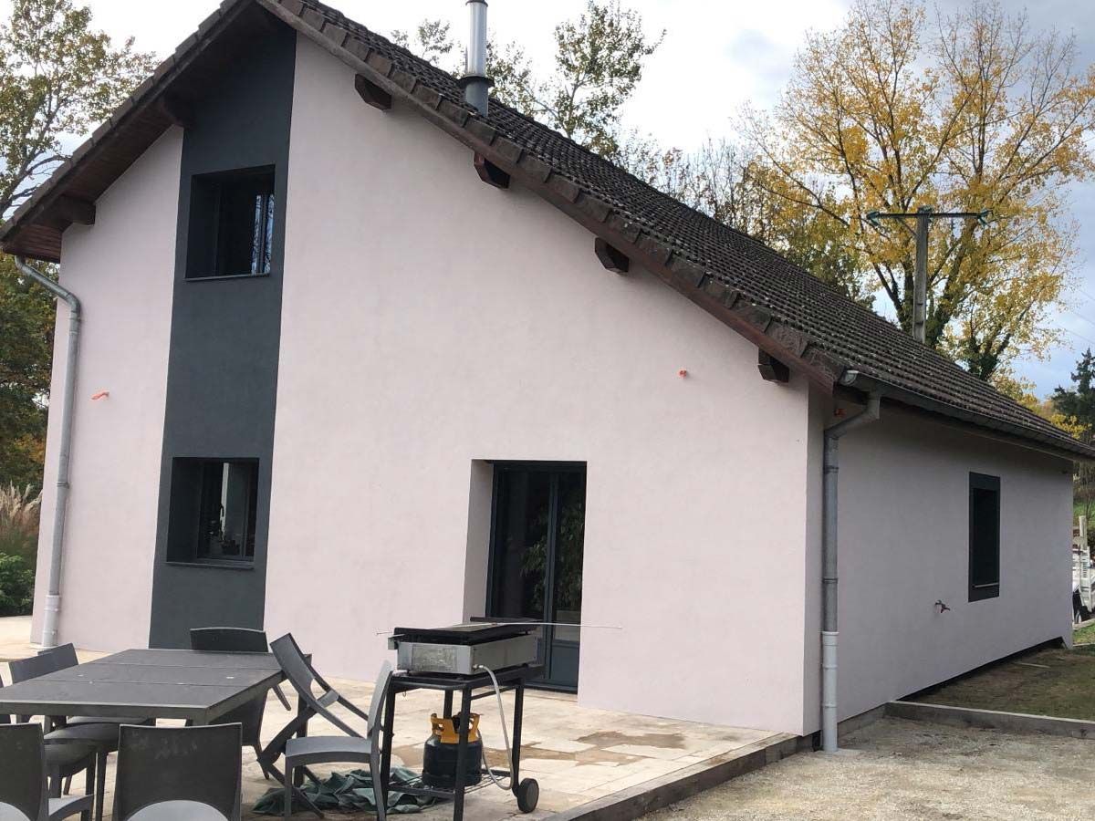 Une maison moderne rose clair avec des finitions gris foncé, un toit à pignon et une terrasse avec un ensemble repas et un barbecue.