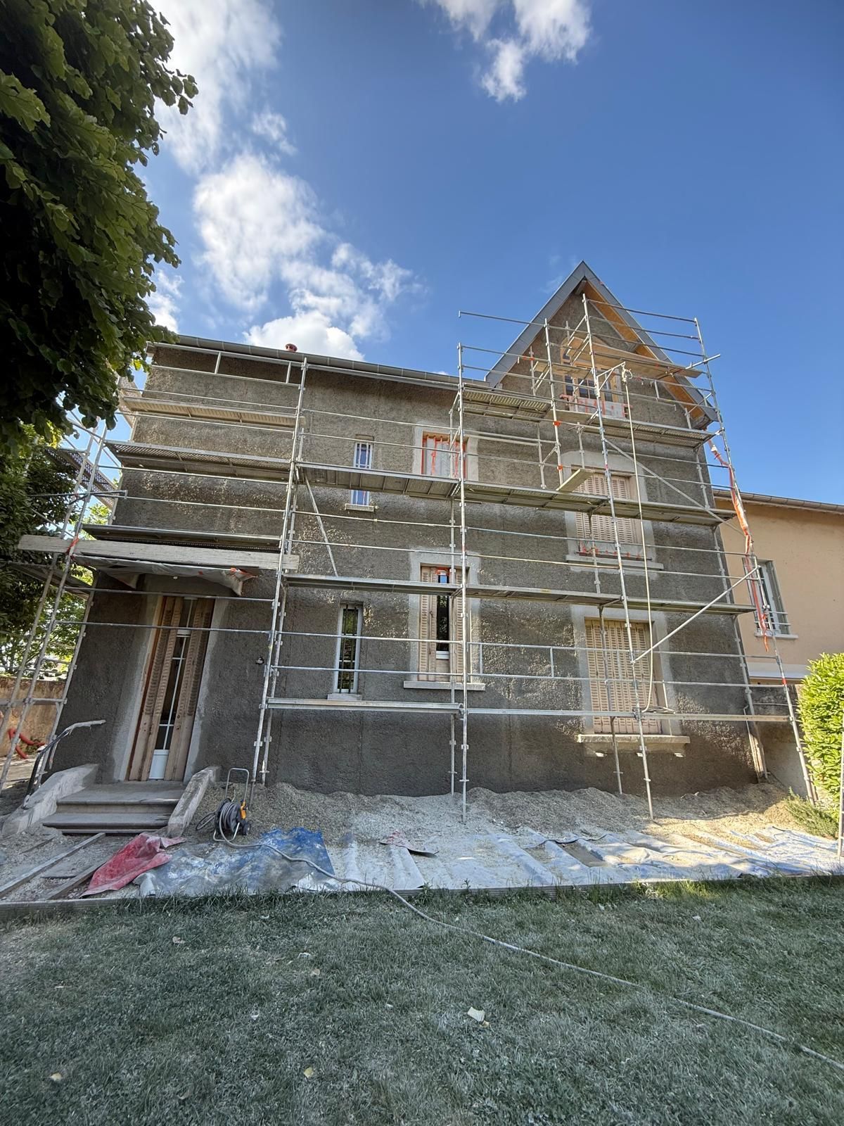 Une maison à plusieurs étages en rénovation, recouverte d'un enduit foncé et entourée d'échafaudages métalliques, par une journée ensoleillée.