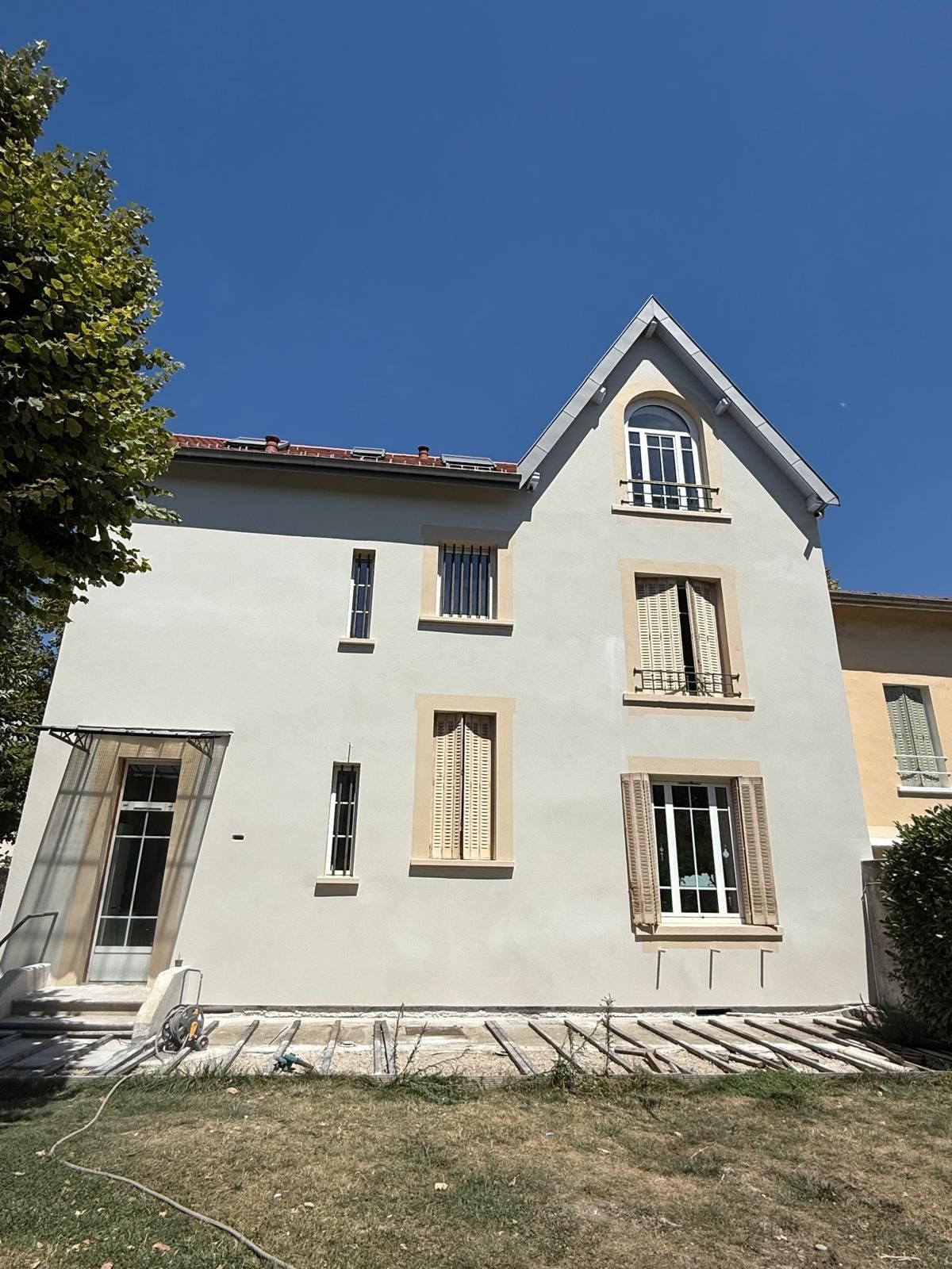 Une maison individuelle beige de trois étages avec un toit pointu, une fenêtre cintrée au sommet et plusieurs fenêtres rectangulaires sous un ciel bleu.