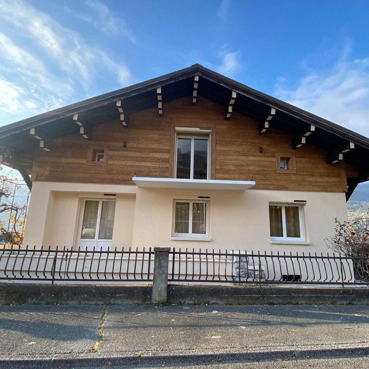 Maison de style chalet en bois avec des murs inférieurs blancs et une cour avant clôturée sous un ciel bleu