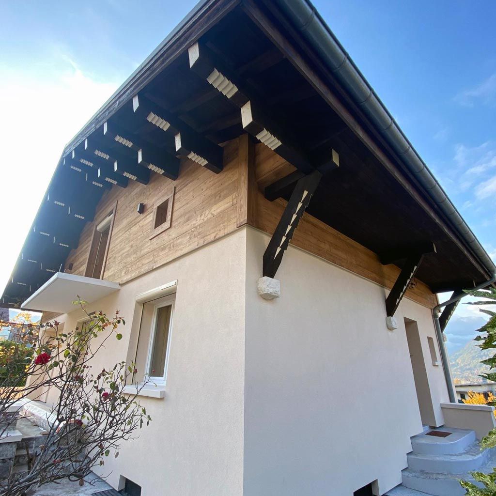 Extérieur d'une maison aux murs blancs, à la façade supérieure en bois et au toit sombre et pentu, sous un ciel bleu.