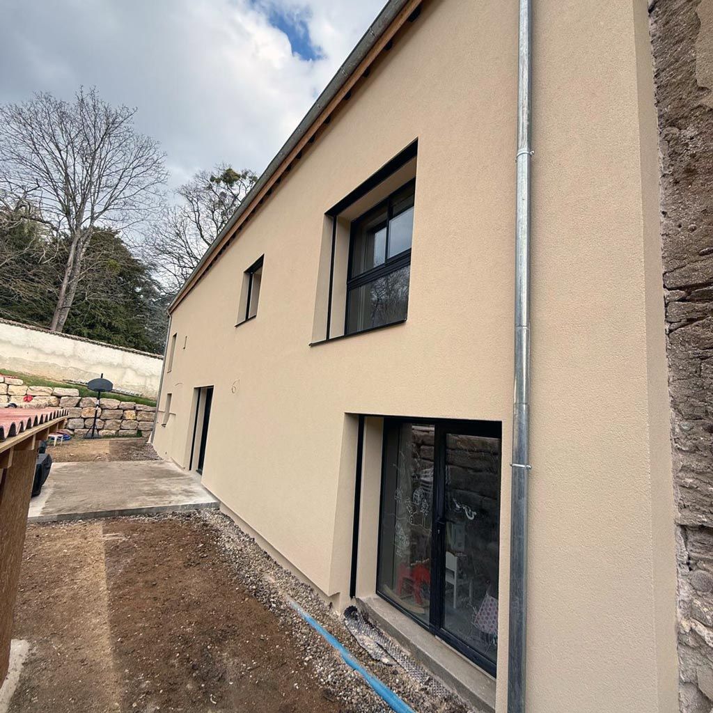 Vue extérieure d'une maison beige en construction, avec un terrain non aménagé et de grandes fenêtres.