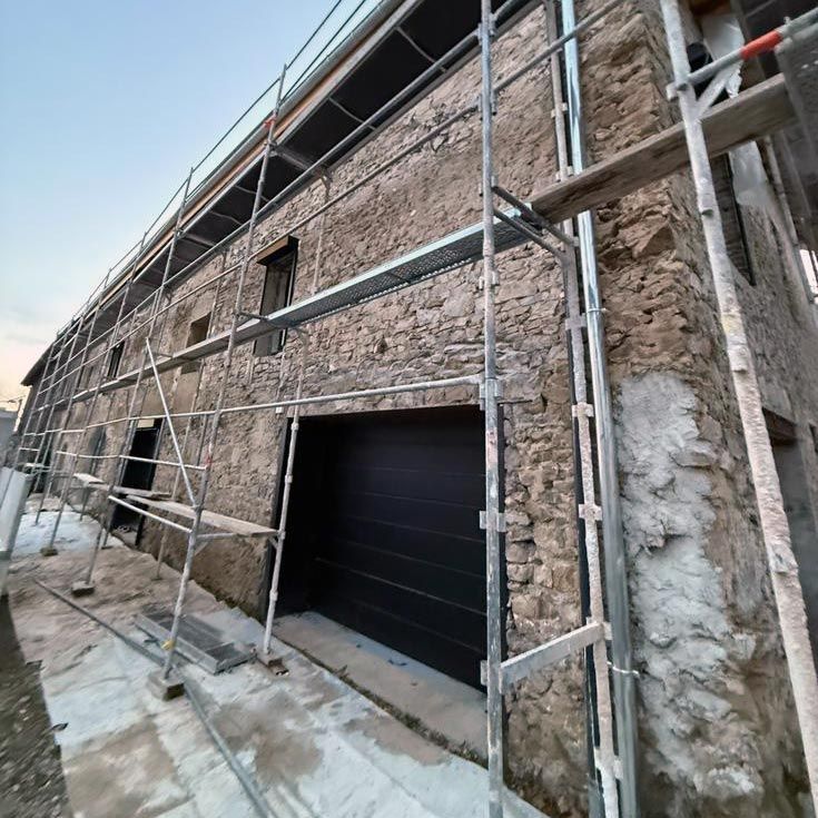 Échafaudages sur la façade en pierre d'un bâtiment en rénovation, avec une grande porte de garage sombre et des murs en plâtre brut.