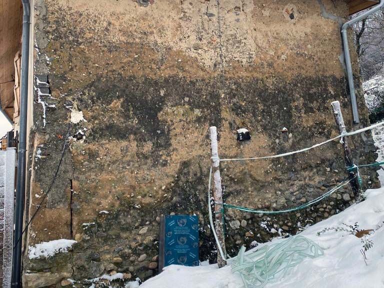 Un mur aux tons terreux, patiné par le temps et reposant sur des fondations en pierre, se dresse à l'extérieur, partiellement recouvert de taches sombres et de plaques de neige.