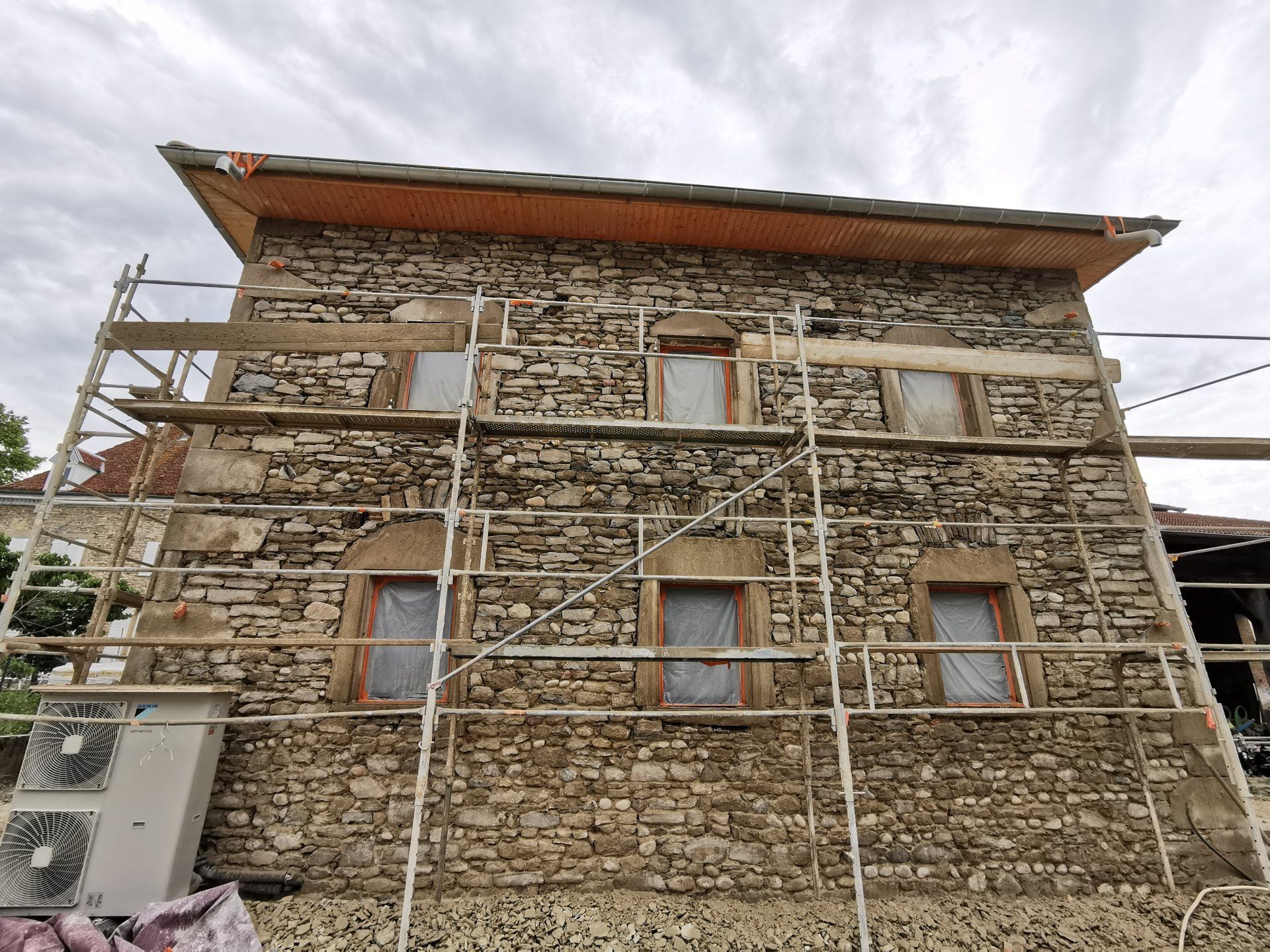 Un bâtiment en pierre en cours de rénovation, dont la façade est recouverte d'échafaudages métalliques, avec des rangées de fenêtres et un avant-toit en bois.