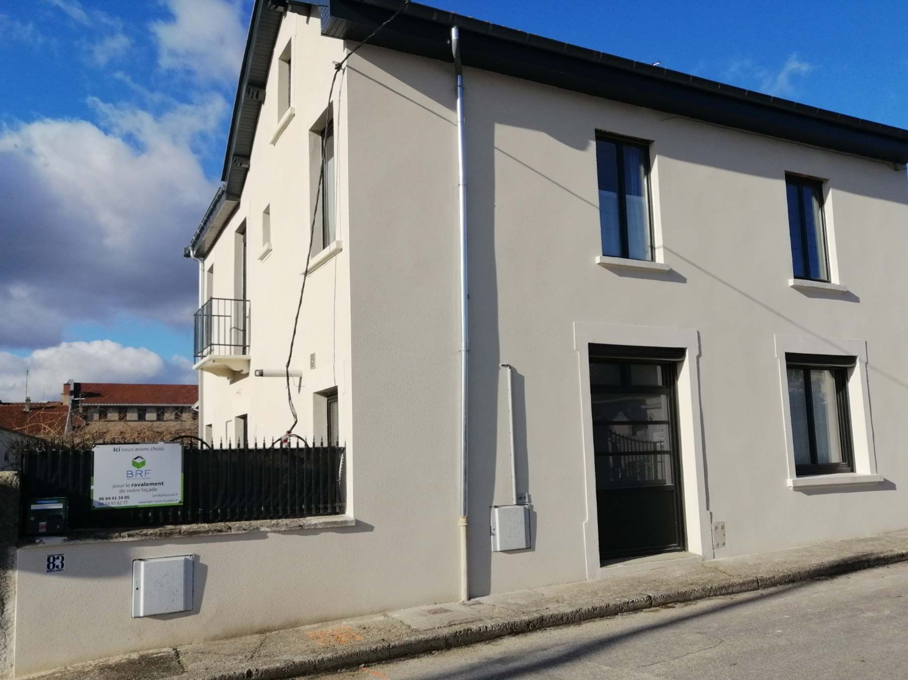 Un bâtiment clair de deux étages avec des fenêtres sombres et une porte noire, doté d'un petit balcon et d'une clôture noire.