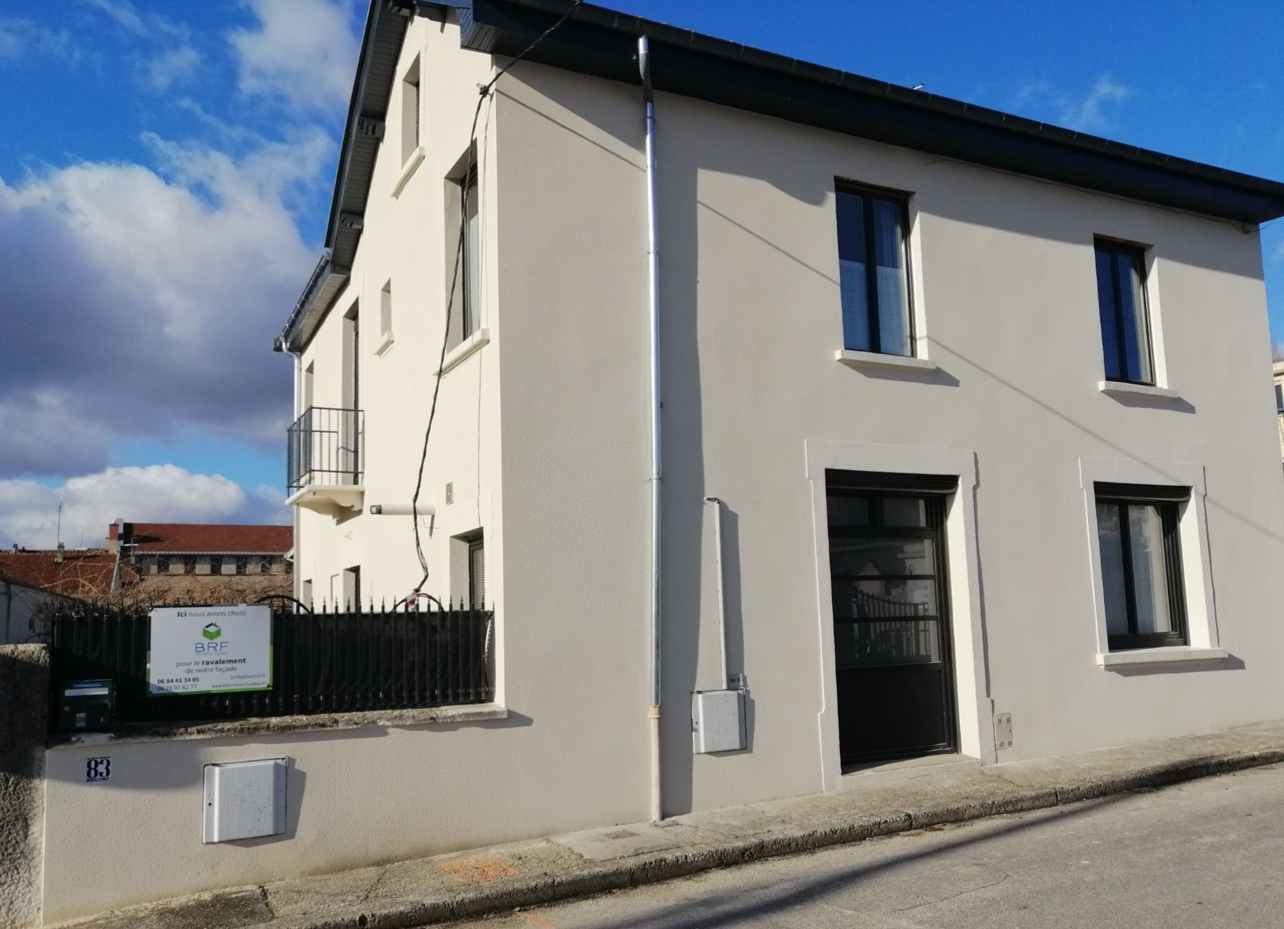 Un immeuble résidentiel de deux étages de couleur claire, avec un toit foncé, des fenêtres à cadre noir et une clôture en métal noir.