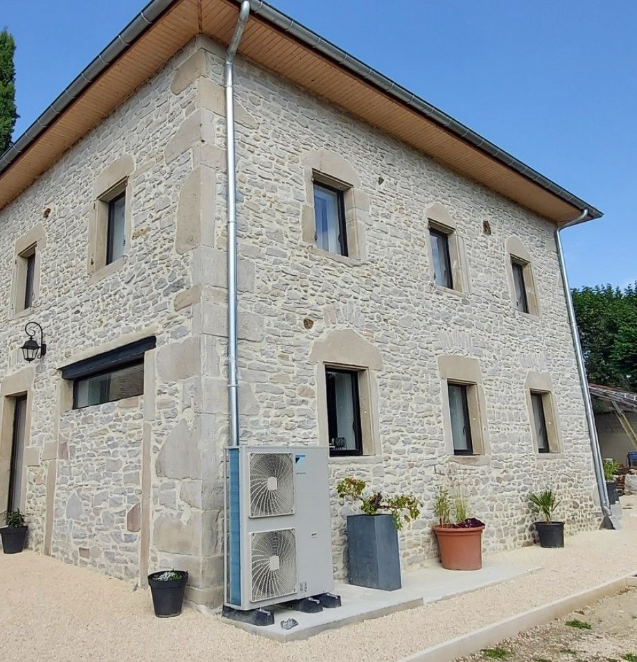 Une maison en pierre sous un ciel bleu azur, avec des fenêtres à cadre sombre, une pompe à chaleur moderne et plusieurs plantes en pot.
