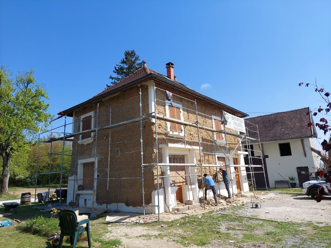 Deux ouvriers, perchés sur des échafaudages, rénovent la façade en briques de terre crue d'une maison à deux étages sous un ciel d'un bleu éclatant.