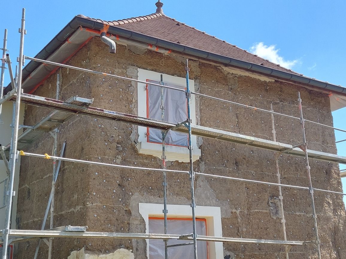 Une maison rustique brune en cours de rénovation, entourée d'échafaudages métalliques avec des ouvertures de fenêtres condamnées.