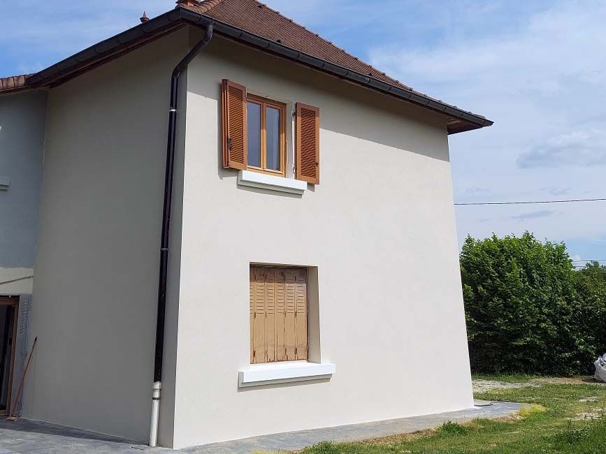 L'extérieur d'une maison claire à deux étages, avec des volets en bois brun à deux fenêtres et un toit de tuiles, se détache sur un ciel bleu.
