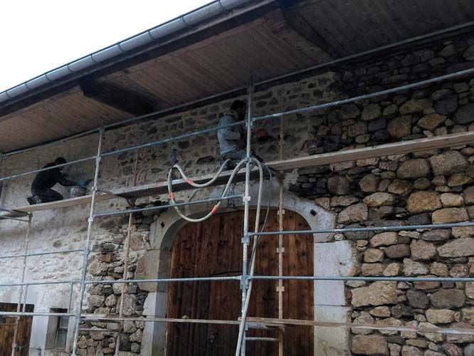 Deux ouvriers, juchés sur un échafaudage, réparent un mur de pierre rustique d'un bâtiment doté d'une grande porte cintrée en bois.