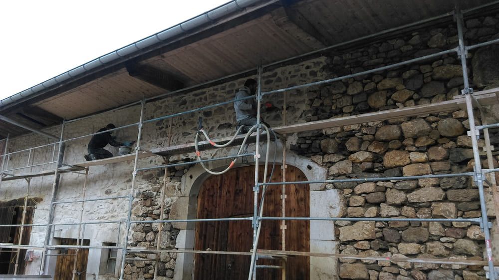 Deux ouvriers, juchés sur un échafaudage, réparent le mur en pierre d'un bâtiment, en plâtrant une partie près d'une porte en bois.