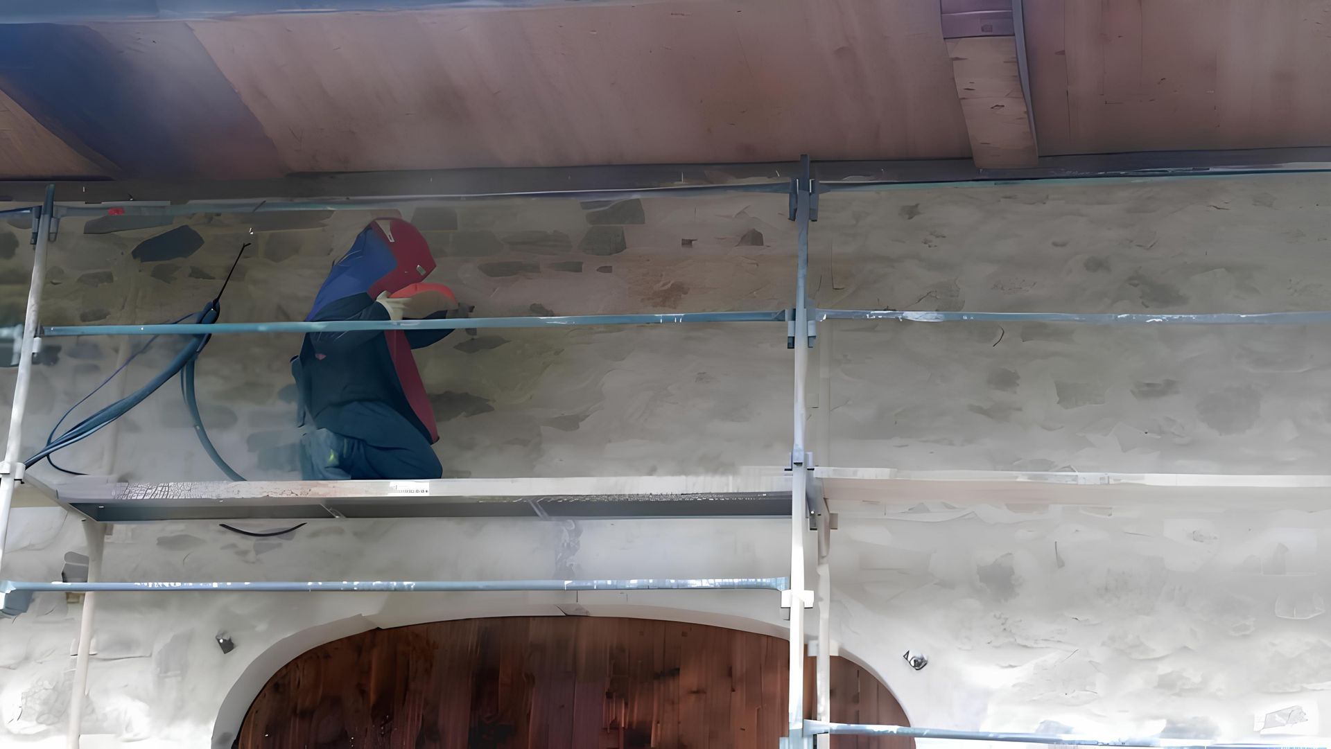 Une personne vêtue de vêtements de travail sombres et coiffée d'une casquette rouge est agenouillée sur un échafaudage pour travailler sur un mur extérieur en pierre.