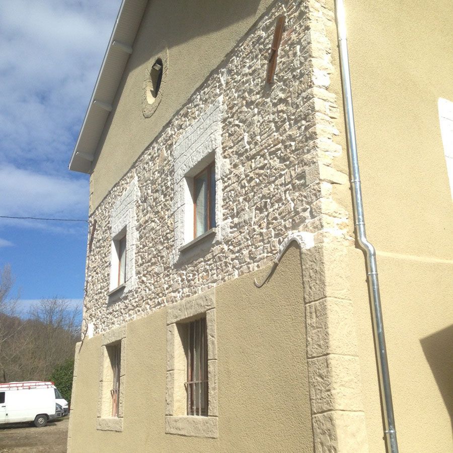 L'extérieur d'un bâtiment de deux étages présente des murs texturés beiges, des éléments en pierre, des fenêtres à cadre brun et une descente de gouttière argentée.