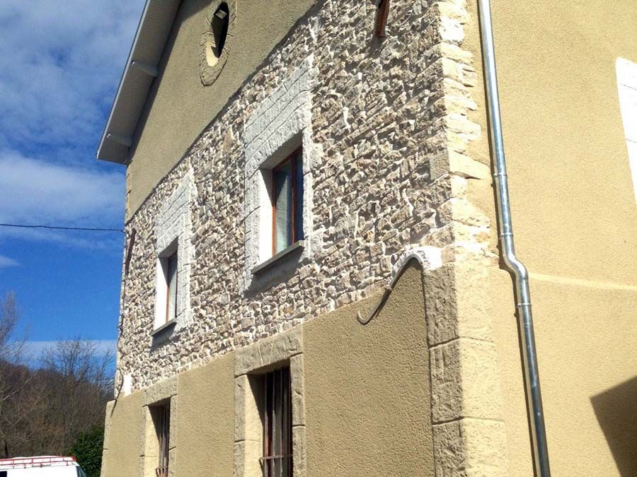 Un bâtiment de deux étages avec une façade en pierre texturée, des ornements en stuc beige et une descente de gouttière en métal, le tout sous un ciel bleu.