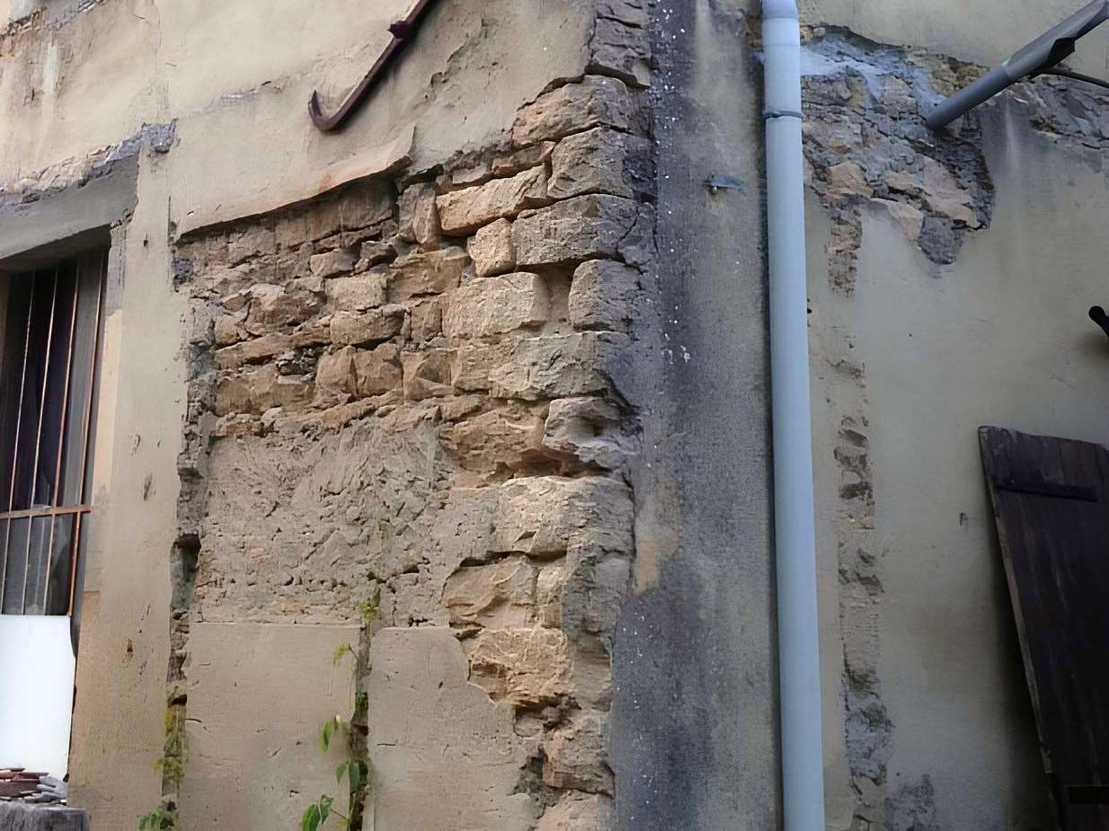 Gros plan sur un mur extérieur endommagé, dont le plâtre s'effrite et laisse apparaître des blocs de pierre clairs et altérés par les intempéries.