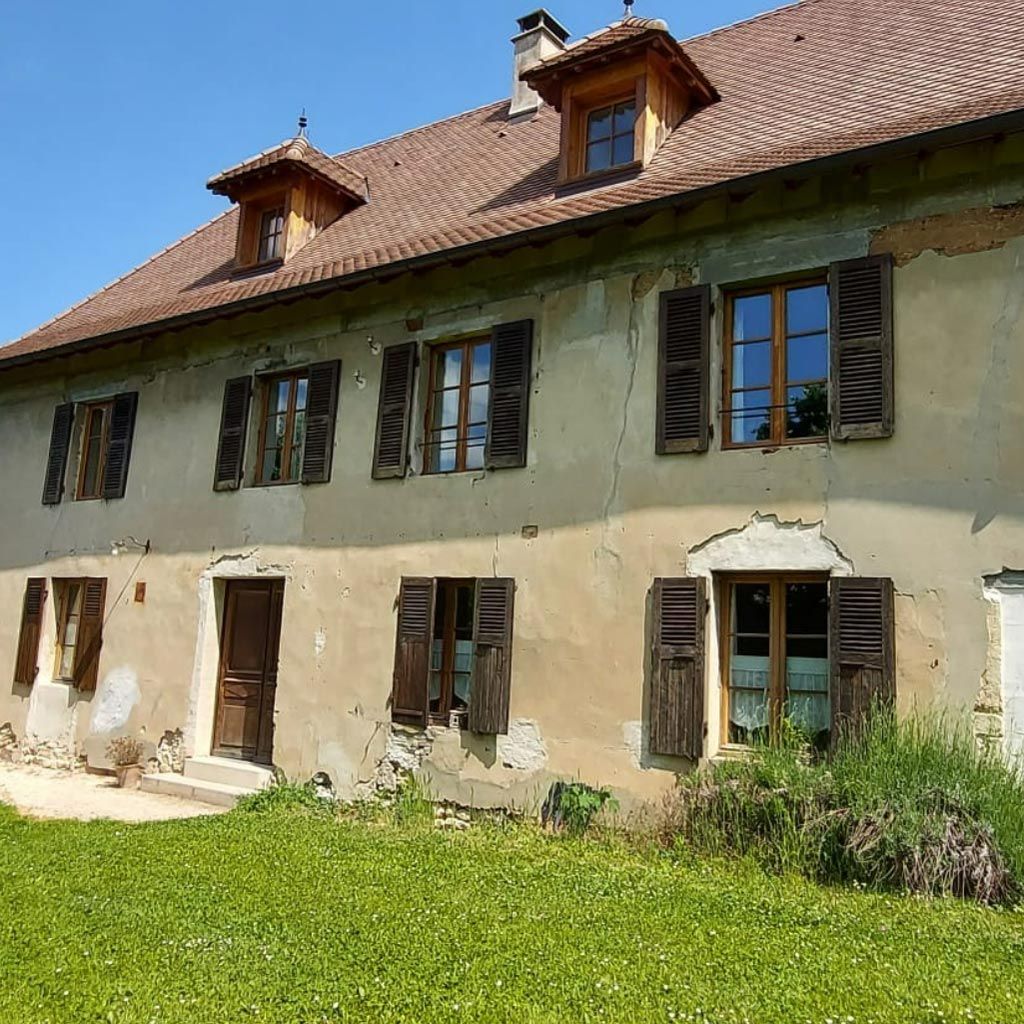Vieille maison rustique aux volets verts, au toit de tuiles rouges et à la cour herbeuse sous un ciel bleu.