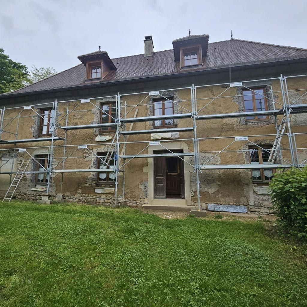 Maison en pierre avec échafaudages en façade, toit marron et pelouse verte devant.
