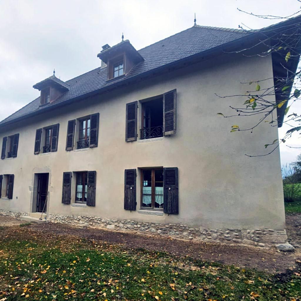 Maison grise à deux étages avec de nombreuses fenêtres et un toit sombre, vue de côté dans une cour arborée