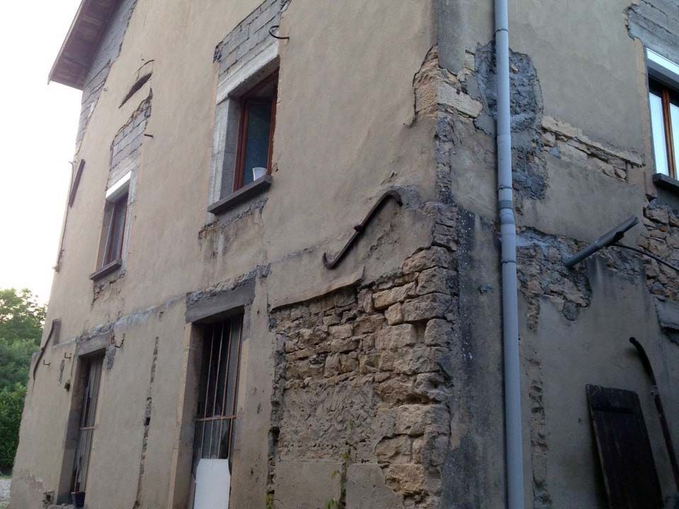 Un bâtiment en pierre de deux étages, usé par le temps, avec un enduit beige abîmé, des maçonneries apparentes près du coin et un tuyau de descente gris.