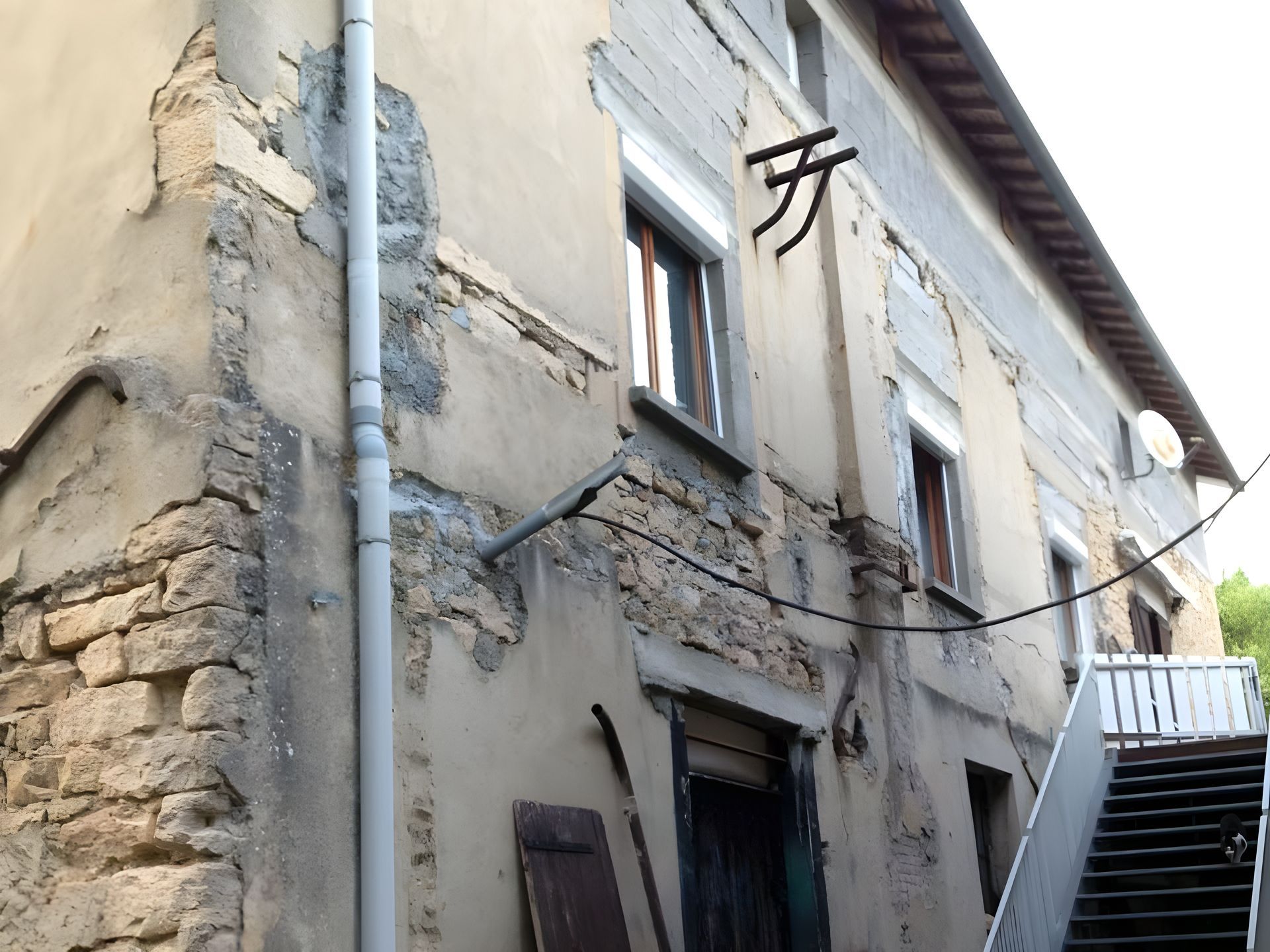 Un vieux bâtiment en pierre délavé par les intempéries, avec un plâtre qui s'écaille, un tuyau de descente gris et un escalier menant à une entrée à l'étage.