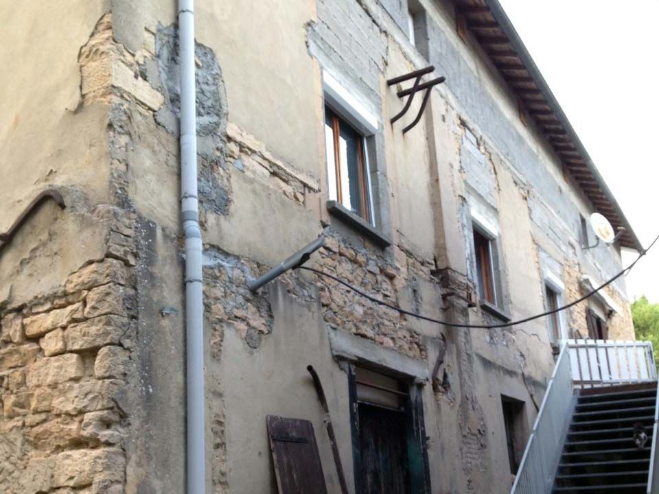 Un vieux bâtiment en pierre délavé par les intempéries, avec un plâtre qui s'écaille, un tuyau de descente gris et un escalier menant à une entrée à l'étage.