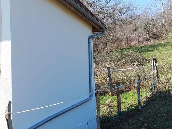 Par une journée ensoleillée, le mur blanc d'une maison, avec sa gouttière grise, longe une clôture en fil de fer qui borde une colline herbeuse.