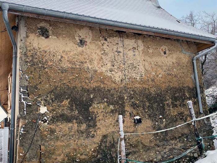 Un mur extérieur en terre ou en pierre, patiné et texturé, présentant des taches sombres et des signes d'érosion sous le bord du toit.