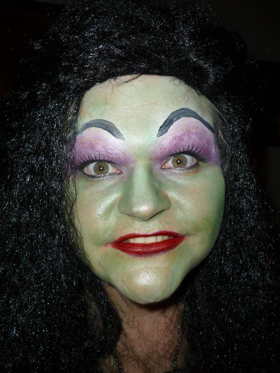 Eine Frau mit grüner Gesichtsbemalung und violetten Augenbrauen trägt eine schwarze Perücke.