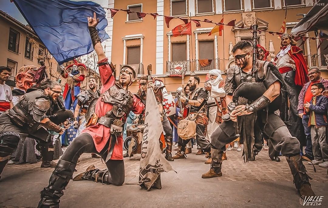 Banda disfrazada tocando tambores y gaitas en medio de una multitud que los anima al aire libre.