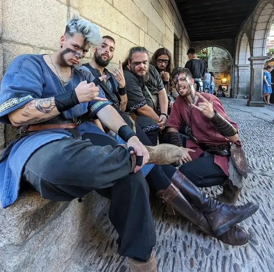 Un grupo de seis personas con trajes medievales posa bajo arcos de piedra. Sonríen, gesticulan y miran a la cámara.