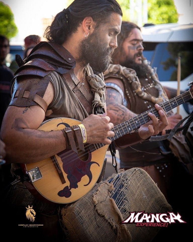 Dos hombres con atuendos medievales tocan instrumentos de cuerda al aire libre. Uno con una mandolina y el otro con un bajo.