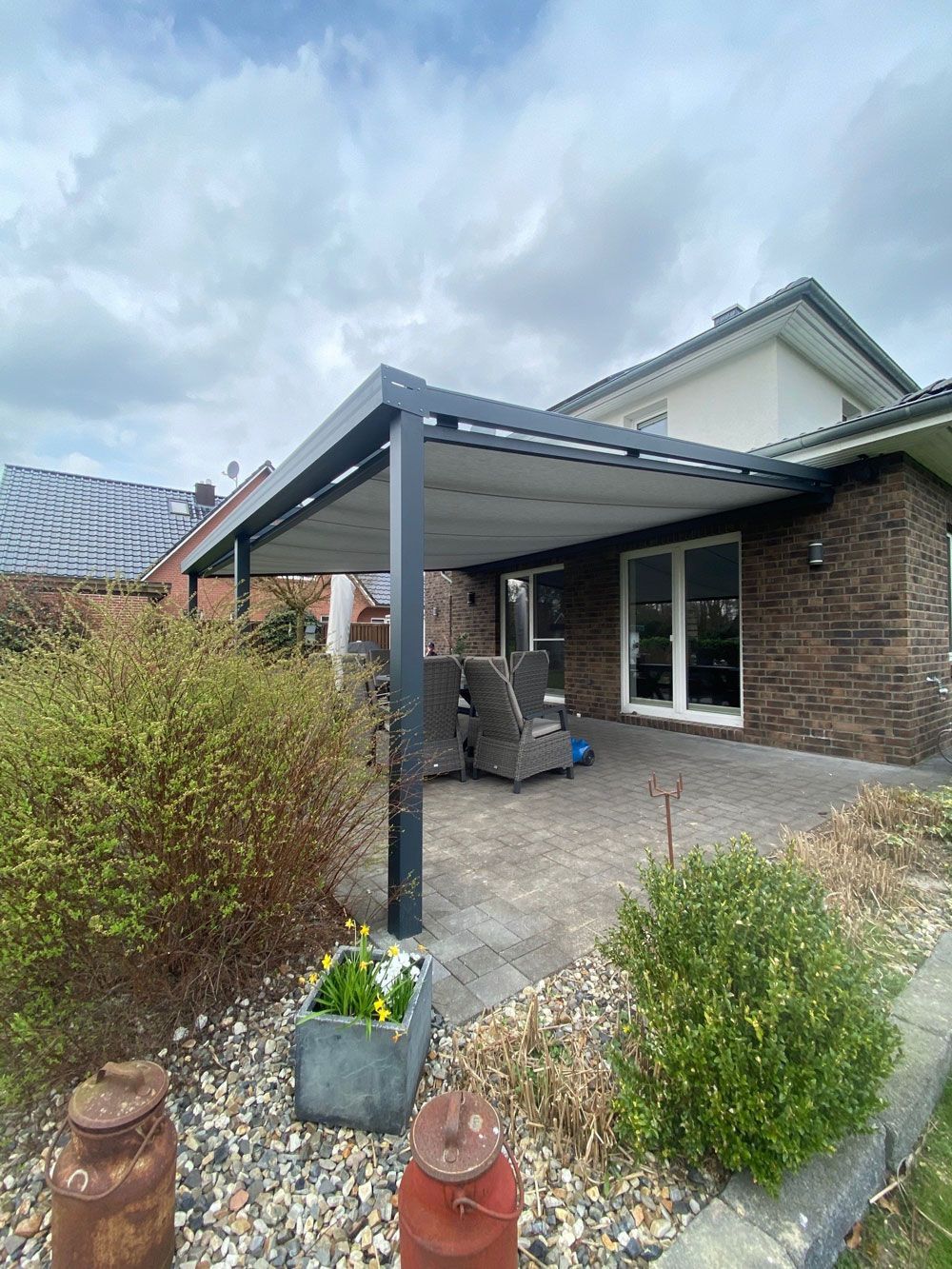 Terrasse mit moderner Pergola, Backsteinhaus, Steinboden und Büschen unter bewölktem Himmel.