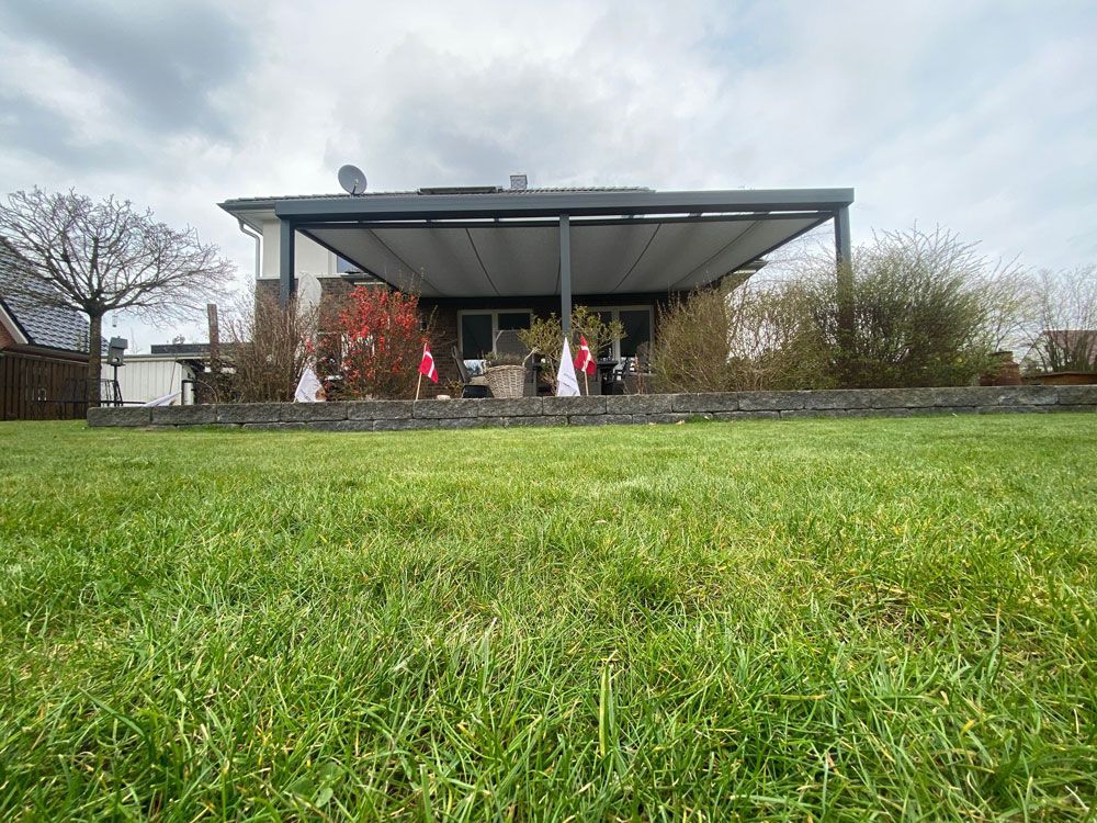 Eine Terrasse im Hinterhof mit ausfahrbarer Markise, Steinmauer und grünem Rasen. Bewölkter Himmel.