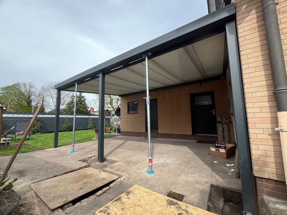 Eine teilweise errichtete graue Pergola ist an ein Backsteingebäude angebaut. Die Stützkonstruktion ist bereits vorhanden; im Vordergrund befindet sich eine Betonplatte.